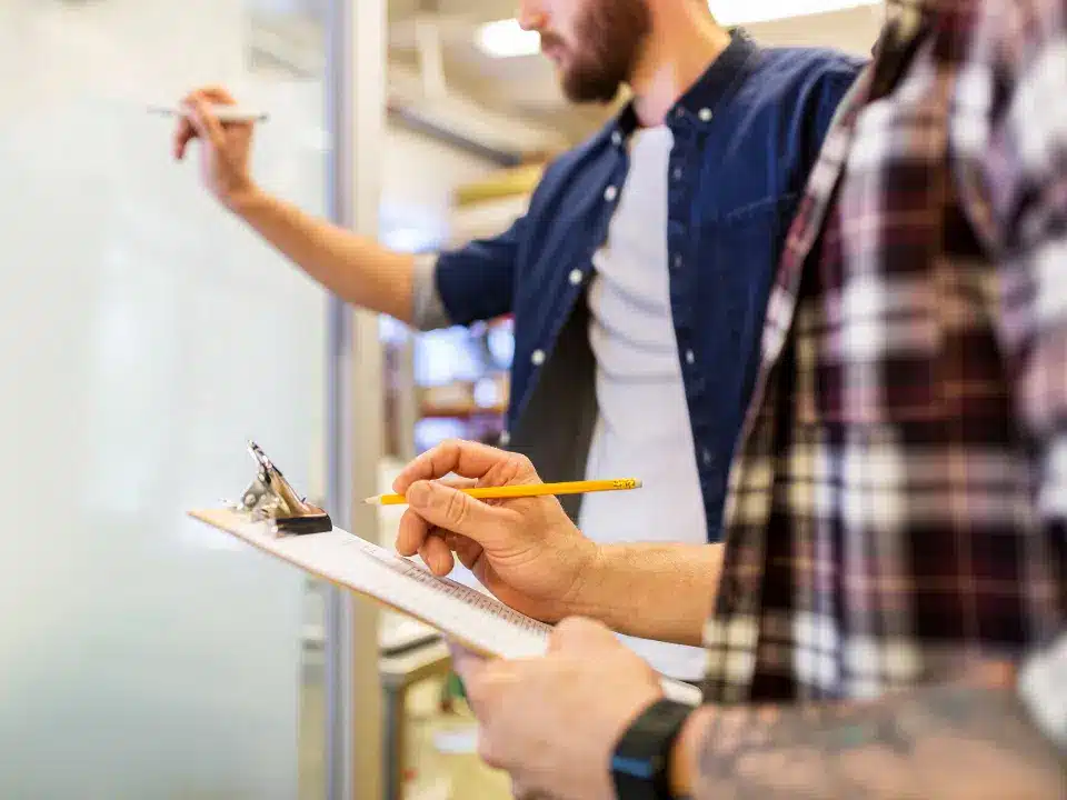Close-up of a person taking notes on a clipboard while a colleague writes on a glass whiteboard in an office.